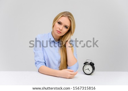Concept cute model misses losing time sitting at the table. Close-up portrait of a beautiful blonde girl with excellent makeup with long smooth hair on a white background in a blue shirt.