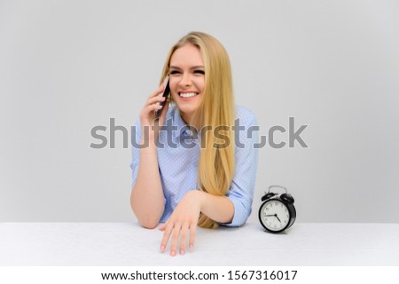 Concept cute model talking on the phone sitting at the table. Close-up portrait of a beautiful blonde girl with excellent makeup with long smooth hair on a white background in a blue shirt.