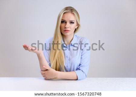 Concept cute model talking to the camera sitting at the table. Close-up portrait of a beautiful blonde girl with excellent makeup with long smooth hair on a white background in a blue shirt.