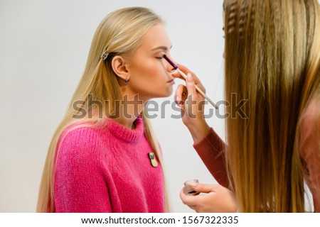 Beauty salon beauty concept. Professional makeup artist with a tool with brushes applies makeup, paints a model with long hair in a pink sweater on a white background. Photo taken in the studio.