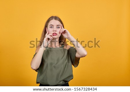 Young beautiful caucasian woman keeping hand on face, model gesture wearing haki shirt on isolated orange background