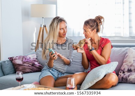 Two female friends gossiping with glasses red wine and pizzas, Showing photo images to her friend on her social media network on the mobile phone