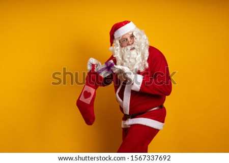 Emotional male actor in a costume of Santa Claus holds a Christmas sock in his hands with gifts and boxes and poses on a yellow background