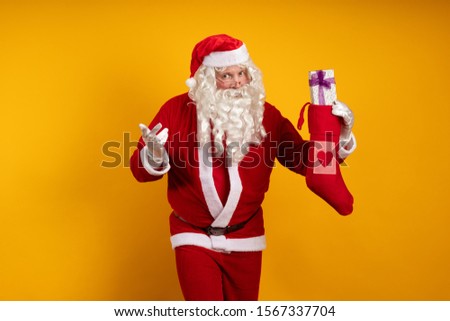 Emotional male actor in a costume of Santa Claus holds a Christmas sock in his hands with gifts and boxes and poses on a yellow background