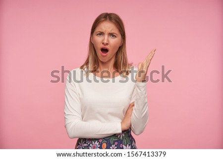 Confused young blonde with casual hairstyle raising palm with displeased face and keeping her mouth opened, wearing white blouse and flowered skirt over pink background