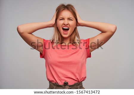 Stressed young pretty blonde female with casual hairstyle holding her head with raised hands, frowning her face with wide mouth opened while looking at camera, isolated over white background