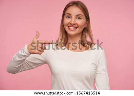 Joyful attractive young blonde woman with casual hairstyle pointing aside with index finger and smiling widely to camera, wearing casual clothes while posing over pink background