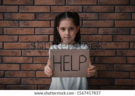 Sad little girl with sign HELP near brick wall. Child in danger