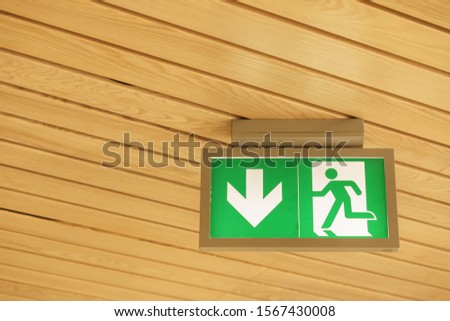 Green alert emergency escape sign lightbox on the wooden ceiling. Icon of a man running away through an open door. A white arrow on a green background shows the downward direction.