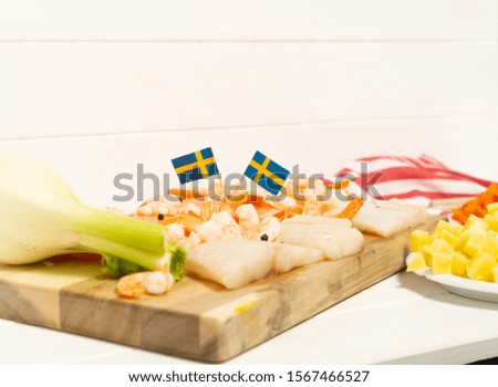 Salmon, cod, fennel on a wooden cutting board. Potatoes and carrots in white plates. Spoon and striped tablecloth. Swedish flags. Ingredients for traditional Scandinavian fish soup. Swedish food.