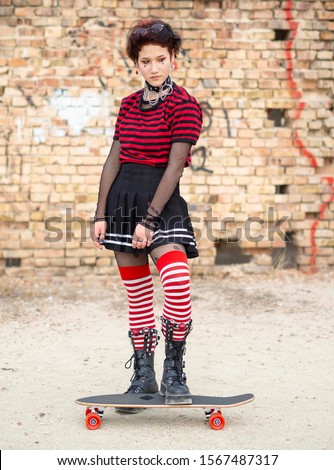 Vertical shot alternative emo girl sitting on a skate board on a brick wall background. Dark teen in a black skirt, striped stocking and steel boots