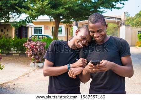 gay couple laughing together while using a phone together. two young black men walking and holding each other laughing together while viewing content on a mobile phone. Royalty-Free Stock Photo #1567557160