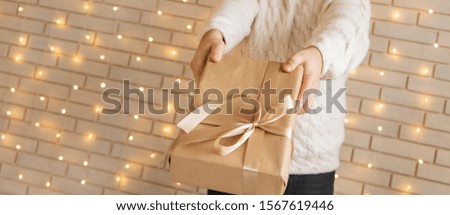 holidays celebration person hands taking gift box wrapped in paper with bow to camera in white interior background brick wall and garland yellow illumination wallpaper pattern  picture with copy space
