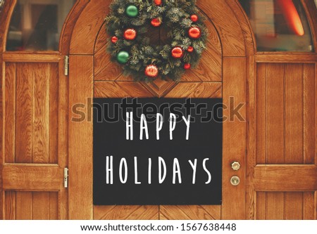 Happy Holidays text sign on  empty board with copy space on rustic wooden doors and stylish christmas wreath with red and green baubles at holiday market. Seasons greeting card