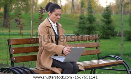 a woman in a business suit and a camel coat turned on a laptop sitting on a wooden bench in the park on a background of greenery. freelance concept