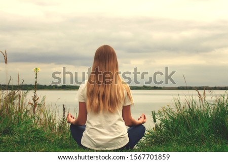 Beautiful girl with long hair is sitting on the shore. The view from the back. Sunset. Peace and tranquility. Yoga by the lake.