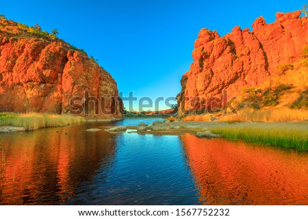 Northern Territory, Australian Outback. Scenic Glen Helen Gorge in West MacDonnell Ranges changes colours with sunrise light and reflects on waterhole in dry season at sunrise light. Royalty-Free Stock Photo #1567752232
