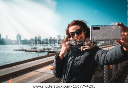 Beautiful curly brunette woman taking selfie self-portrait with the Brookling bridge on the background while sightseeing new york during winter season