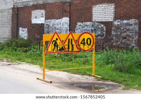 Group of yellow road signs (repair work, narrowing of the road and speed limit)  on the side of a road. Green grass and old brick wall on the background