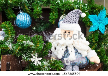 soft toy figurine of Santa Claus under Christmas tree with snow, snowflakes, garlands and blue balls on wooden background. Traditional home decor, symbol for Christmas, New Year holidays.