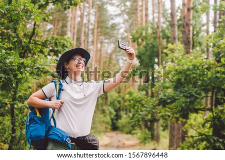 Smiling female tourist holds smart phone, takes selfie against beautiful landscape wood. Hiking woman with backpack taking selfie photo with smartphone. Travel and healthy lifestyle outdoors.