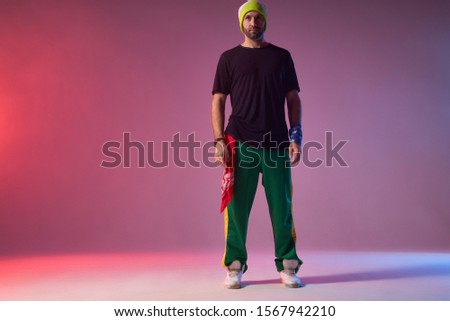 Handsome tall man with thick stylish beard dressed in casual sportive clothes, ready to start training in dance studio, teaching hip hop dance, waiting for people gathered, light coloured background