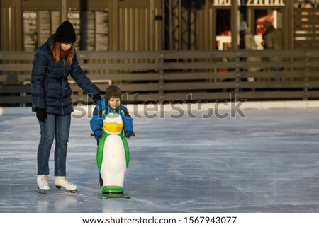 The boy is skating at the rink. The child learns with his parent to ice skate. A kid is standing in a blue jacket amid a crowd of people.