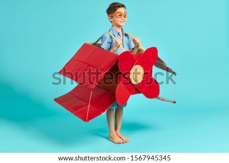 Little caucasian boy flying on red carboard airplane, happy boy in casual clothes imagine himself as pilot. Isolated blue background, sky