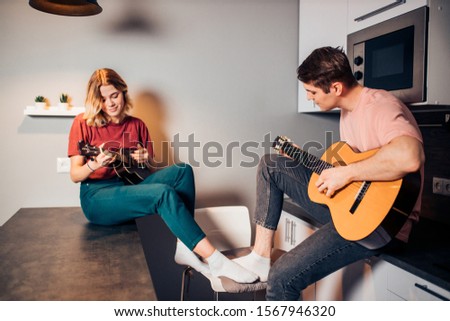 handsome self-taught boy playing guitar while her girlfriend playing ukulele isolated in kitchen.