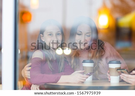 Mother and daughter sitting in cafe drinking coffee. One girl looking on the camera. woman looking on mobile phone. Picture through window.