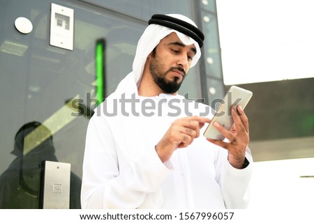 Arab Emirati man scrolling his smart mobile phone with modern business background ideal for corporate mockup. Middle East local male wearing kandura dish dash