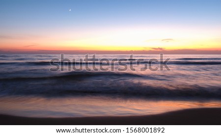 The sun is setting. Amazing sunset over Mediterranean sea in Italy. Clear and colorful evening sky with the white moon. Beautiful landscape with diffuse reflection on water. Long exposure of wavy sea.