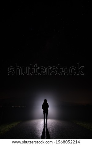Silhouette of a woman in the darkness. Night Photography. Bright light shining behind dark mysterious figure. Ghostly, mystical, surreal person standing. Royalty-Free Stock Photo #1568052214