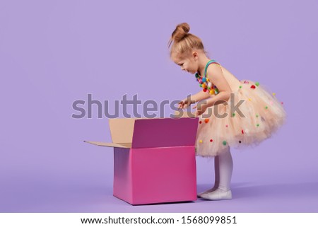 A funny little girl child in a magnificent holiday dress look inside a large gift colored box, on a purple isolated background. Birthday surprise