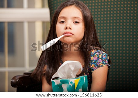 Little girl suffering from a cold, holding a box of tissues and a thermometer in her mouth