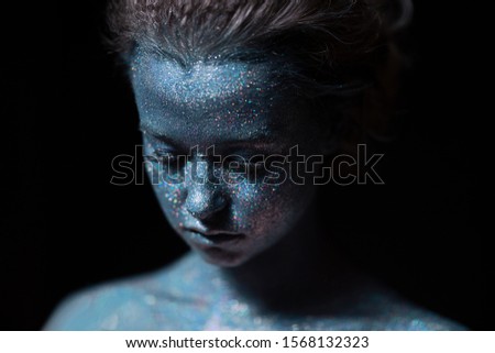 girl with a blue face in profile in sparkles close-up on a black wall looks down