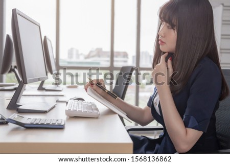 Beautiful Asian woman wearing a dark blue suit sitting're taking notes in a notebook at a desk in a modern office and a willingness to work and a large glass window background.