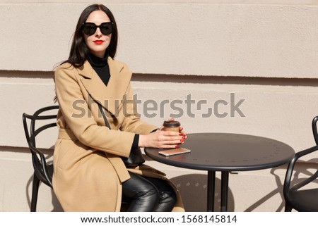 Horizontal picture of brunette tender slender young woman, holding her hands around papercup of coffee, having smartphone on table surface, wearing black accessories, lunchtime. Relax concept.
