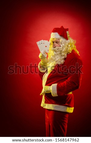 Male actor in a costume of Santa Claus holds playing cards in his hands and poses on a dark red background