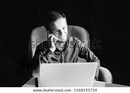 young guy sitting in armchair and talking on the phone on black and white photo. laptop on the table in front of a guy.