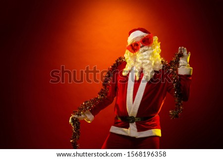 Male actor in a costume of Santa Claus in large pink glasses and with a red garland of tinsel in his hands dancing and posing on a dark red background