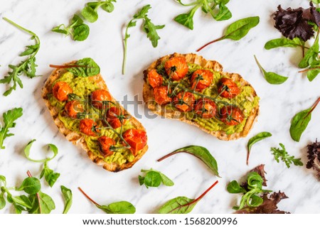 Toasts with mashed avocado guacamole and oven dried cherry tomatoes. The two toasts are seen from above flat lay perspective and there are salad leaves scattered on the table.