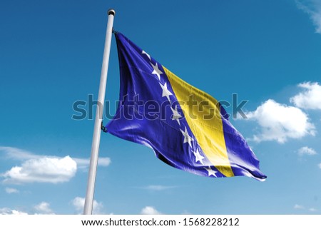 Waving Flag of Bosnia in Blue Sky. Bosnia Flag on pole for Independence day. The symbol of the state on wavy cotton fabric. Royalty-Free Stock Photo #1568228212