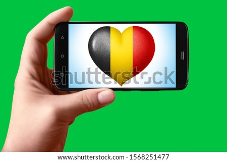 Belgium flag in a heart shape on the screen. Smartphone in hand shows the heart of the flag on the background hromakey. Object for installation and design.