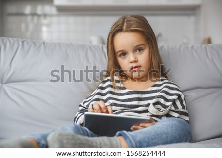 Little child girl indoors sitting on the coach looking camera watching cartoons on digital tablet leisure