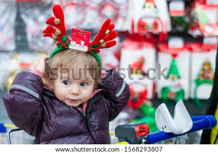 Cute little girl trying on the rim with deer antlers and bells on the background of Christmas decoration. Sitting in the shopping cart. Holiday shopping.