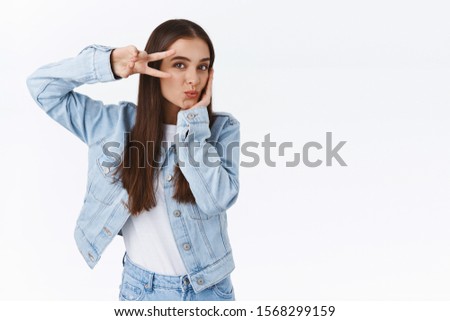 Coquettish, feminine good-looking brunette woman in denim outfit, showing peace or victory sign over eye, tilt head and pouting flirty, smiling as making tender, cute grimaces, white background