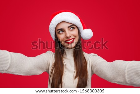 happy young female in Santa hat looking away and dreaming while taking selfie during Christmas party against red background