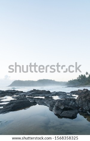 A long exposure shot of a cloudy sunrise from Resende Beach in Itacare, Bahia, Brazil. The white water rock pools are in front of the surf bay with sea spray mist covering the mountains beyond.