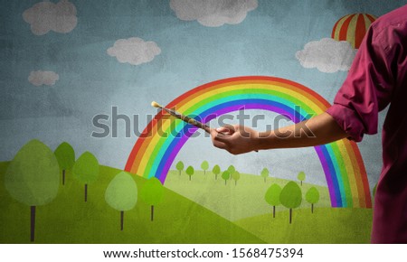 Close up artist hand holding paintbrush. Painter in shirt standing on background colorful picture. Summer landscape with green field, blue sky and rainbow artwork. Creative hobby and profession.
